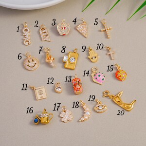 May include: A selection of gold-coloured charms featuring diverse designs such as an apple, cross, smiley face, passport, and a shark. Some charms are adorned with crystals and colourful enamel.