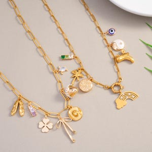 May include: Gold-toned charm necklaces with various pendants, including a four-leaf clover, ballet slippers, a bow, a heart, a rainbow, and a palm tree. The necklaces have a delicate chain link design and are displayed on a neutral background.