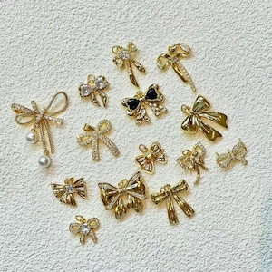 May include: A selection of gold-toned bow-shaped charms and embellishments. Some bows are decorated with clear crystals, while others have black details or hanging pearls. The charms are in various sizes and designs, ideal for jewellery or craft projects.