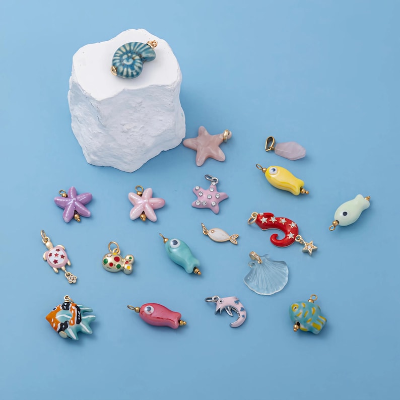 May include: A collection of colorful, sea-themed charms, including starfish, fish, seahorses, and shells. The charms are in various colors like pink, blue, yellow, and red, with gold-colored accents. They are arranged on a light blue background.