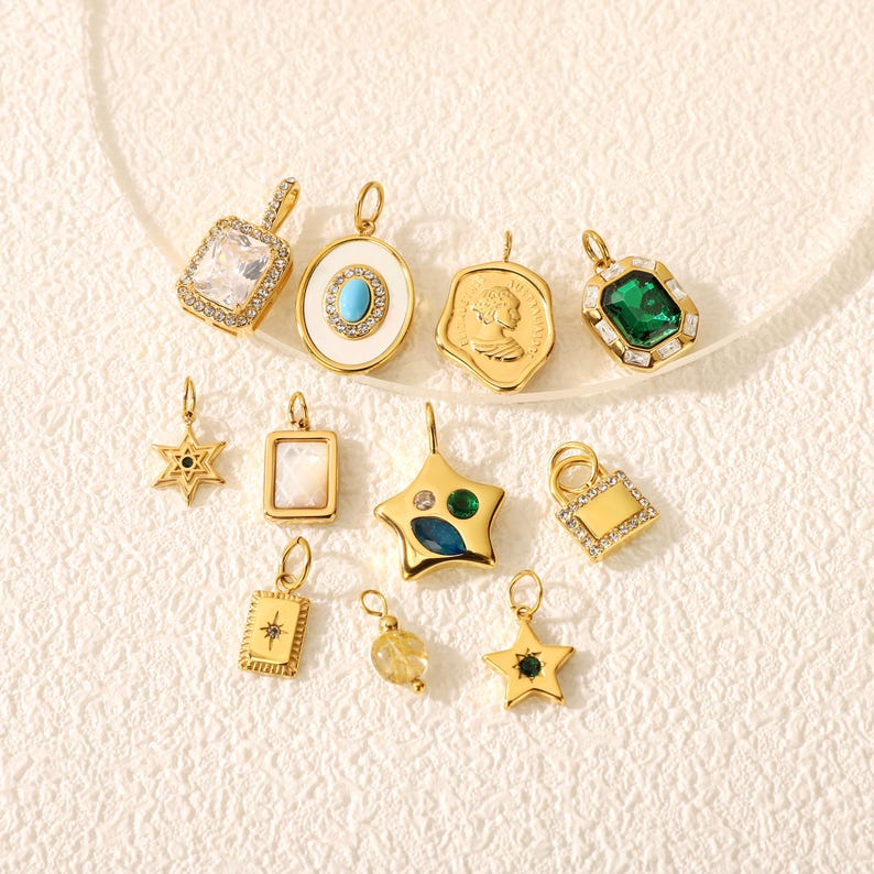 May include: A collection of gold-toned pendants in various shapes, including stars, squares, and ovals. Some pendants feature gemstones in green, blue, and clear, while others have intricate designs. The pendants are displayed on a light-colored surface.
