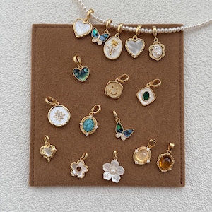 Peut inclure: Une collection de breloques en métal doré aux formes variées, notamment des cœurs, des fleurs, des papillons et une étoile, certaines incrustées de nacre, de turquoise ou de pierres ambrées, disposées sur une carte de présentation carrée marron.