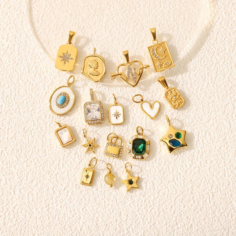 May include: A collection of gold-colored pendants with various designs, including stars, hearts, and geometric shapes. Some pendants feature gemstones, while others have intricate patterns. The pendants are displayed on a textured, off-white surface.