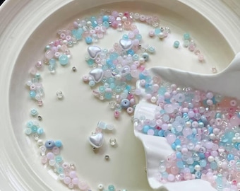 May include: A collection of pastel-colored beads, including pink, blue, and clear, are scattered on a white plate and in a seashell-shaped dish. Some beads are heart-shaped. The beads vary in size and texture, creating a delicate and whimsical aesthetic.