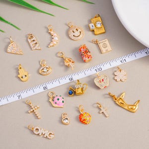 May include: A selection of gold-coloured charms featuring diverse designs such as a smiley face, cross, heart, and passport. Some charms are adorned with clear crystals. A ruler is placed across the charms.