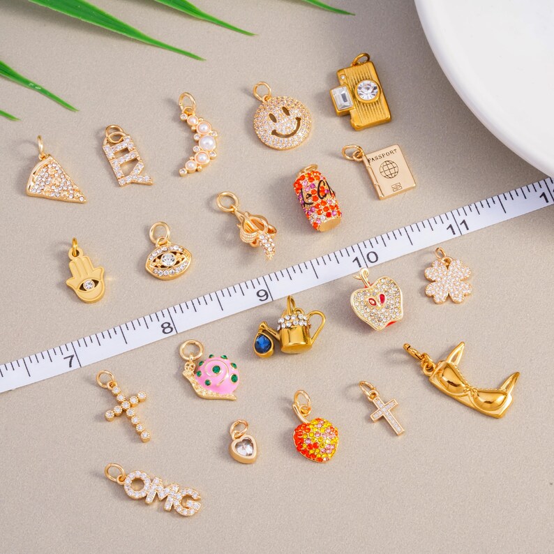 May include: A selection of gold-coloured charms featuring diverse designs such as a smiley face, cross, heart, and passport. Some charms are adorned with clear crystals. A ruler is placed across the charms.
