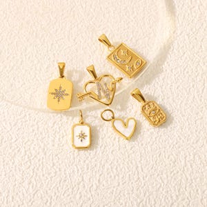 May include: A collection of gold-colored pendants with various designs. The pendants include a starburst, a heart pierced by an arrow with the letter "N", a rectangle with a crescent moon, an eye, and a heart-shaped pendant with a white inlay.
