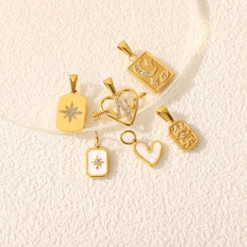 May include: A collection of gold-colored pendants with various designs. The pendants include a starburst, a heart pierced by an arrow with the letter "N", a rectangle with a crescent moon, an eye, and a heart-shaped pendant with a white inlay.