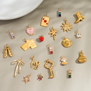 May include: A selection of gold-coloured charms in diverse shapes, such as a heart, a playing card, a strawberry, a sun, a key, and a bow. Some charms feature colourful gemstones. The charms are displayed on a light grey surface.