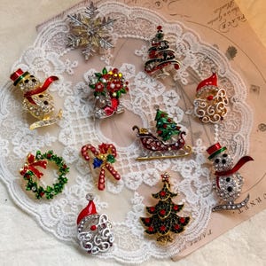 Puede incluir: Una colección de diez broches de metal con temática navideña que incluye un copo de nieve plateado, dos muñecos de nieve con bufandas rojas, dos caras de Papá Noel, dos árboles de Navidad decorados, una corona, un trineo y un bastón de caramelo.