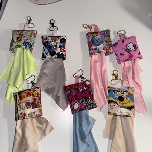 May include: Seven square fabric pouches with metal lobster clasps, each containing a microfiber cleaning cloth. The pouches feature various colorful cartoon character prints, including Hello Kitty, Stitch, and Disney princesses, secured with snap buttons.