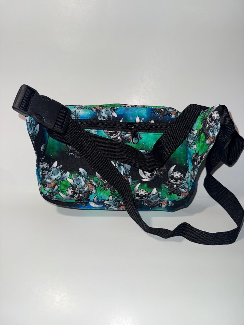 May include: A black fanny pack with a colorful pattern featuring a cartoon character. The bag has a black adjustable strap and a black buckle closure. The background of the pattern is a mix of blue and green.