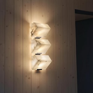 Könnte beinhalten: Moderne Wandleuchte mit drei geometrischen, würfelförmigen Leuchten. Die Leuchten bestehen aus einem durchscheinenden, marmorähnlichen Material, das ein warmes Licht ausstrahlt. Montiert an einer hellen Holzwand.