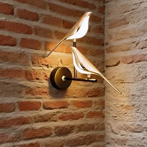 May include: A wall-mounted light fixture featuring two bird-shaped lamps. The lamps are silver-colored with a sleek design, mounted on a black arm and a circular base. The fixture is set against a brick wall.