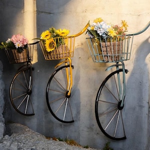 Op de afbeelding: Drie vintage-stijl fietsen met zwarte metalen frames en wielen zijn aan een muur gemonteerd. Elke fiets heeft een draadmand gevuld met kleurrijke bloemen. De manden zijn geel, groen en blauw.