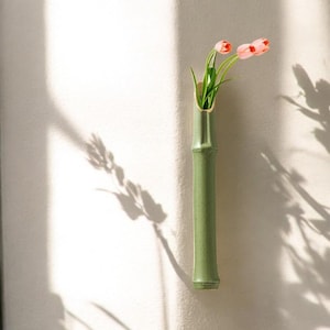 May include: A green bamboo vase with a small opening at the top, holding pink tulips. The vase is mounted on a white wall.