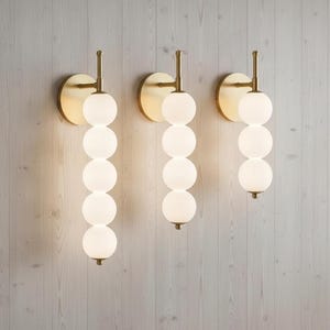May include: Three wall sconces with a brass finish and a series of five white, spherical glass bulbs. The sconces are mounted on a light-colored wooden wall, creating a modern and elegant lighting fixture.
