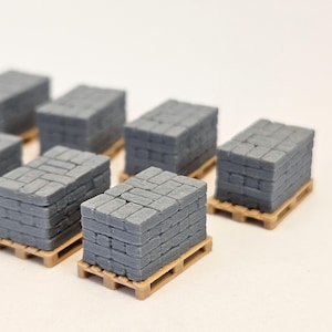 May include: Miniature gray stone bricks stacked on small wooden pallets. The bricks have a textured surface, and the pallets are a light brown color. Several stacks are visible, arranged in a close-up shot.