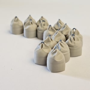 Puede incluir: Una colección de pequeños objetos en forma de saco, de color gris claro, con una parte superior fruncida y un pequeño acento azul. Los objetos están dispuestos sobre una superficie blanca, algunos en primer plano y otros en segundo plano.