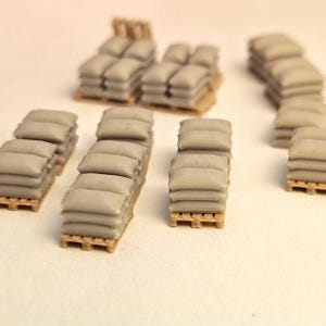 Puede incluir: Miniaturas de pilas de sacos de arena de color gris claro, dispuestas sobre pequeñas paletas de madera. Se observan varias pilas, algunas con cuatro capas y otras con cinco, lo que sugiere un tema de construcción o militar. Las paletas son de madera.