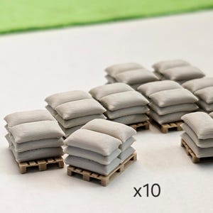 Op de afbeelding: Miniatuur stapels grijze zandzakken op kleine houten pallets. De afbeelding toont meerdere stapels, met de tekst "x10" die de hoeveelheid aangeeft. De zandzakken zijn neutraal grijs van kleur en de pallets zijn lichtbruin.