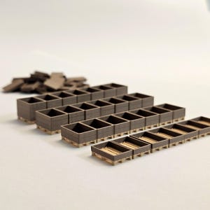May include: A collection of small, brown wooden crates, arranged in rows, with a pile of similar pieces in the background. The crates have a textured appearance and are set on light-colored pallets. The image is well-lit, with a focus on the miniature crates.