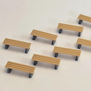 May include: A collection of miniature wooden benches. Each bench has a light brown slatted seat and dark grey legs. The benches are arranged on a white surface, likely for a model railway or similar project. The benches are approximately 5 cm in length.