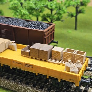 May include: A yellow model train flatcar loaded with miniature cargo, including wooden crates, stacks of lumber, and burlap sacks. A brown train car filled with coal is in the background. The scene is set on a model railroad track.