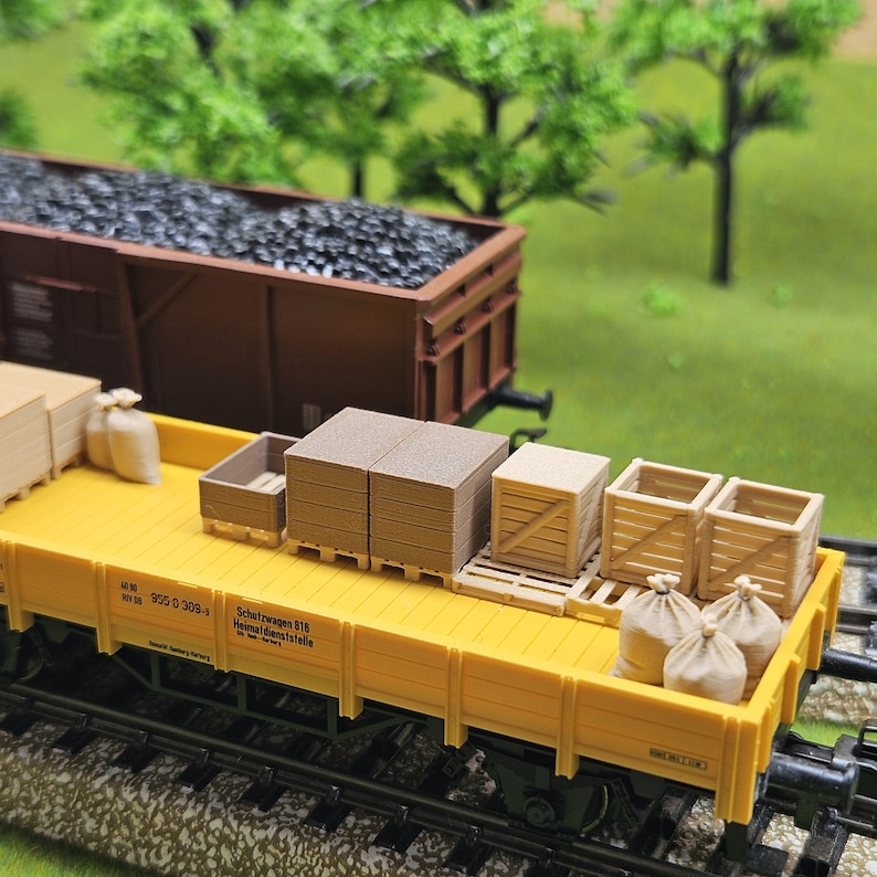 May include: A yellow model train flatcar loaded with miniature cargo, including wooden crates, stacks of lumber, and burlap sacks. A brown train car filled with coal is in the background. The scene is set on a model railroad track.