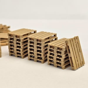 May include: A collection of miniature wooden pallets in a light brown hue. The pallets are stacked neatly, with some leaning against the stacks. The image is well-lit, showcasing the texture and detail of the wood.