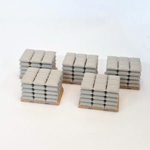 May include: Five miniature pallets stacked with grey, square-shaped bags. Each pallet holds a grid of these small, uniform items arranged in multiple layers, creating a structured, industrial appearance with a neutral color palette throughout.