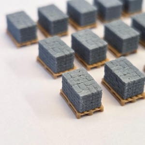 May include: Miniature grey stone blocks stacked on small wooden pallets. The blocks have a textured surface, and the pallets are a light brown colour. Multiple stacks are visible, arranged on a white surface.