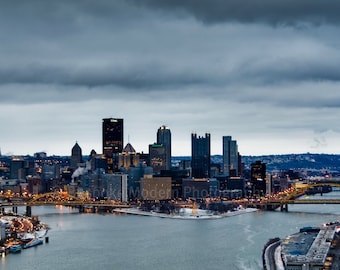 May include: A panoramic view of a city skyline at dusk with a river in the foreground. The city is lit up with lights and there are several tall buildings. The sky is overcast with dark clouds.