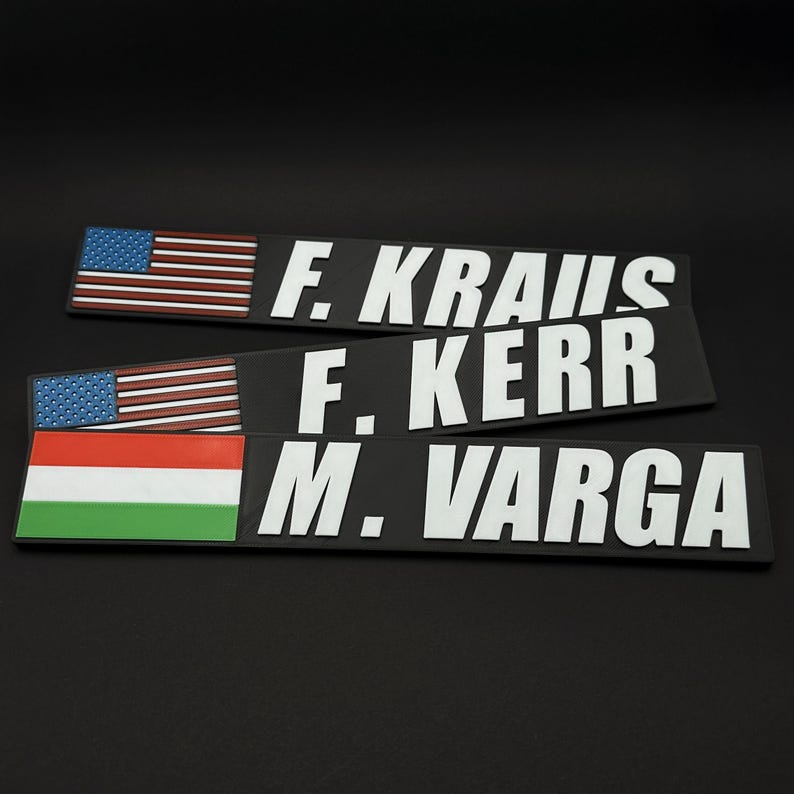 May include: Three black nameplates with white lettering. Two nameplates have the American flag, and one has the Hungarian flag. The names on the plates are F. KRAUS, F. KERR, and M. VARGA.