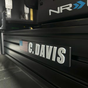 May include: Black metal plate with the text "C. DAVIS" in white, and a small American flag on the left side. The plate is mounted on a black frame, possibly part of a racing simulator setup.