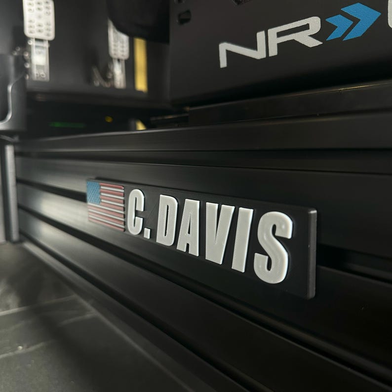 May include: Black metal plate with the text "C. DAVIS" in white, and a small American flag on the left side. The plate is mounted on a black frame, possibly part of a racing simulator setup.