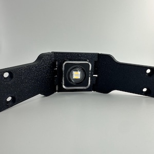 May include: A black plastic light fixture with a white LED light in the center. The light is mounted on a black bracket that can be attached to a surface.