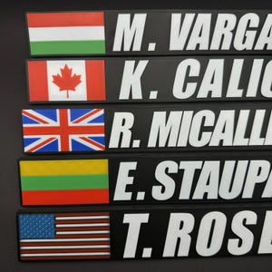 May include: A close-up of five black nameplates with white text and flags. The flags include Hungary, Canada, the United Kingdom, Lithuania, and the United States. The names are M. VARGA, K. CALIC, R. MICALLI, E. STAUP, and T. ROSE.