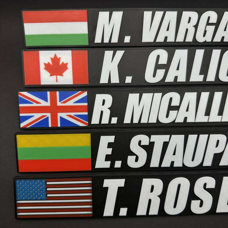 May include: A close-up of five black nameplates with white text and flags. The flags include Hungary, Canada, the United Kingdom, Lithuania, and the United States. The names are M. VARGA, K. CALIC, R. MICALLI, E. STAUP, and T. ROSE.