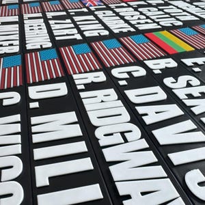 May include: A collection of black rectangular plaques with white raised lettering and colorful flags. The flags include the American flag, the British flag, and the Lithuanian flag. The names are in white, and the overall design is a 3D effect.