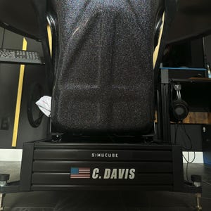 May include: A black racing simulator cockpit with a black seat and a black metal frame. The frame has the text "SIMUCUBE" and "C. DAVIS" with an American flag. The seat is black with yellow accents.