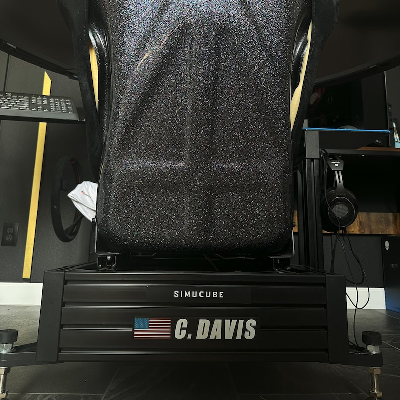 May include: A black racing simulator cockpit with a black seat and a black metal frame. The frame has the text "SIMUCUBE" and "C. DAVIS" with an American flag. The seat is black with yellow accents.