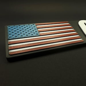 May include: A rectangular emblem featuring the American flag design. The flag has a blue field with white stars, red and white stripes. The emblem is 3D printed with a textured surface and a black border.