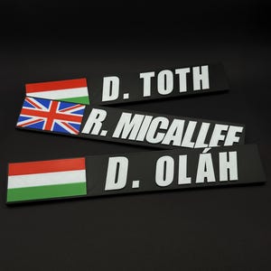May include: Three black nameplates with white text and flags. One nameplate has the Hungarian flag, another the British flag, and the third the Hungarian flag. The names are D. TOTH, R. MICALLEF, and D. OL&Aacute;H.