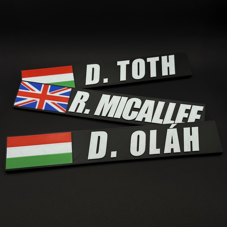 May include: Three black nameplates with white text and flags. One nameplate has the Hungarian flag, another the British flag, and the third the Hungarian flag. The names are D. TOTH, R. MICALLEF, and D. OL&Aacute;H.