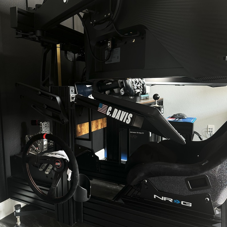May include: A black racing simulator setup featuring a steering wheel, seat, and monitor. The setup includes the text "C. DAVIS" and "NRG". The steering wheel has red accents.