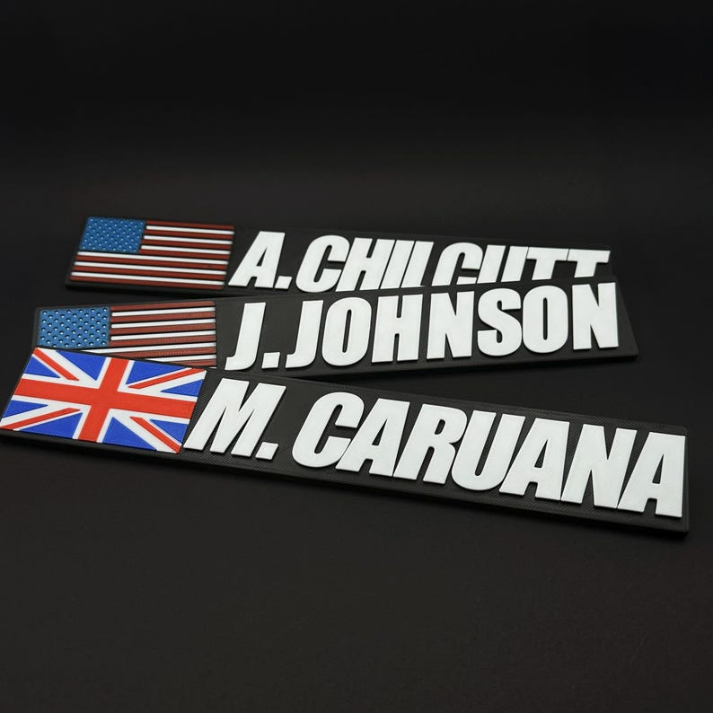 May include: Three black nameplates with white lettering and flags. Two nameplates have the American flag, and one has the British flag. The names are A. CHILCUTT, J. JOHNSON, and M. CARUANA.