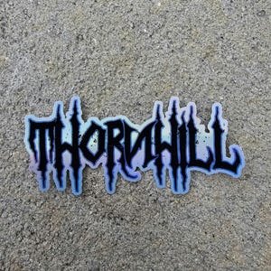 May include: A sticker with the word "THORNHILL" in a gothic font. The text is black with a blue and iridescent outline. The sticker is on a textured gray surface.