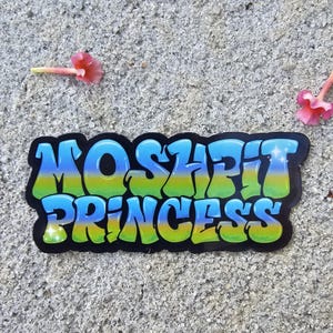 May include: A sticker with the words "MOSHEIT PRINCESS" in a blue, green, and yellow gradient, outlined in black. The sticker is on a textured gray surface, with pink flowers nearby.