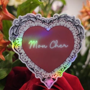 May include: A heart-shaped sticker with the words "Mon Cher" in a cursive font. The sticker has a red center and a decorative white and black border. The sticker is placed in front of a red rose and other colorful flowers.